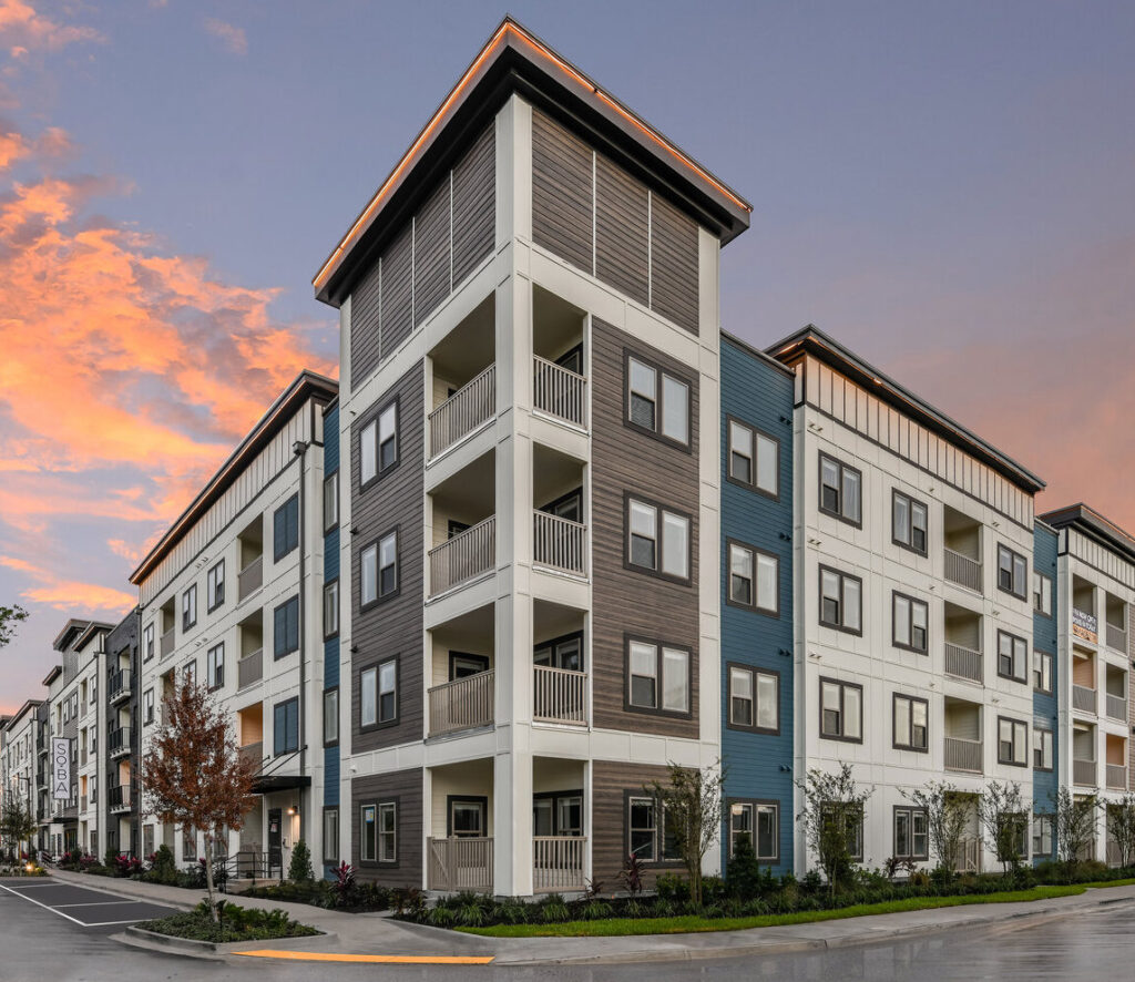 Modern multi-story apartment complex featuring a mix of white, blue, and wood-toned siding with private balconies and large windows. The building is well-maintained and landscaped, set against a vibrant sunset sky.