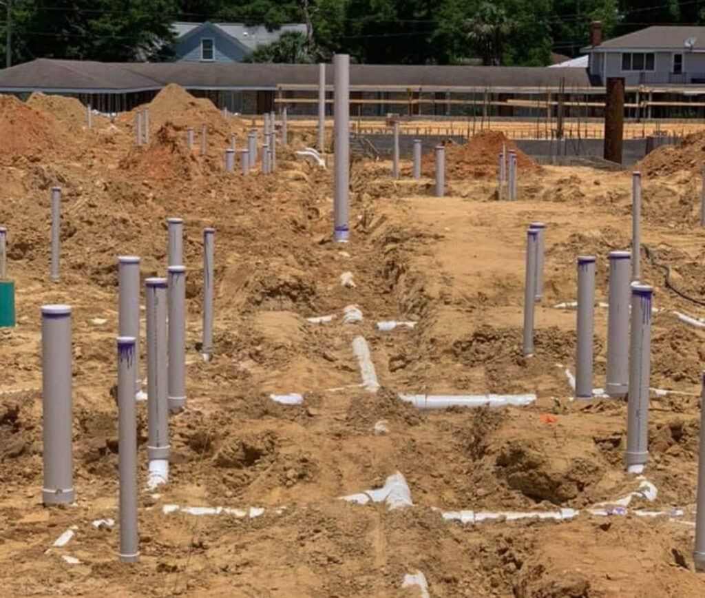 Close-up view of a construction site showing exposed underground plumbing with numerous white PVC pipes emerging from freshly dug trenches. The pipes are capped with purple primer, indicating preparation for a future concrete foundation pour.
