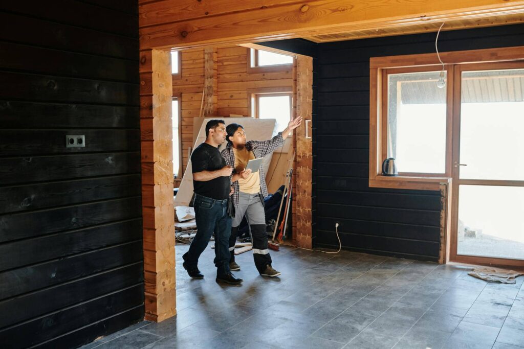 Two men inside a partially finished wooden cabin discuss renovation plans, with one holding a tablet and gesturing toward the ceiling or wall. The room features exposed wood beams, dark paneling, and scattered construction materials, suggesting active remodeling work.