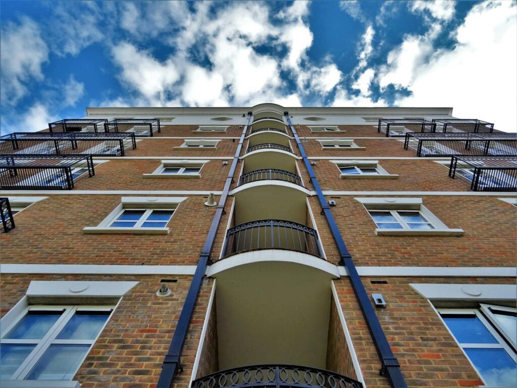 Low-angle view of a multi-story brick apartment building with white trim, black balcony railings, and a central column of curved balconies extending upward. The building is set against a vibrant blue sky with scattered clouds, emphasizing its height and symmetry.