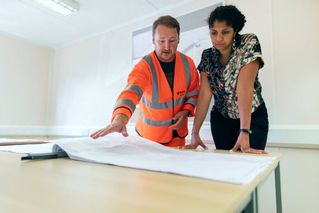 A construction project manager in a high-visibility orange jacket gestures toward a set of blueprints on a table while explaining plans to a colleague. The woman, dressed in a patterned blouse, leans in to closely review the architectural drawings in a well-lit office space.